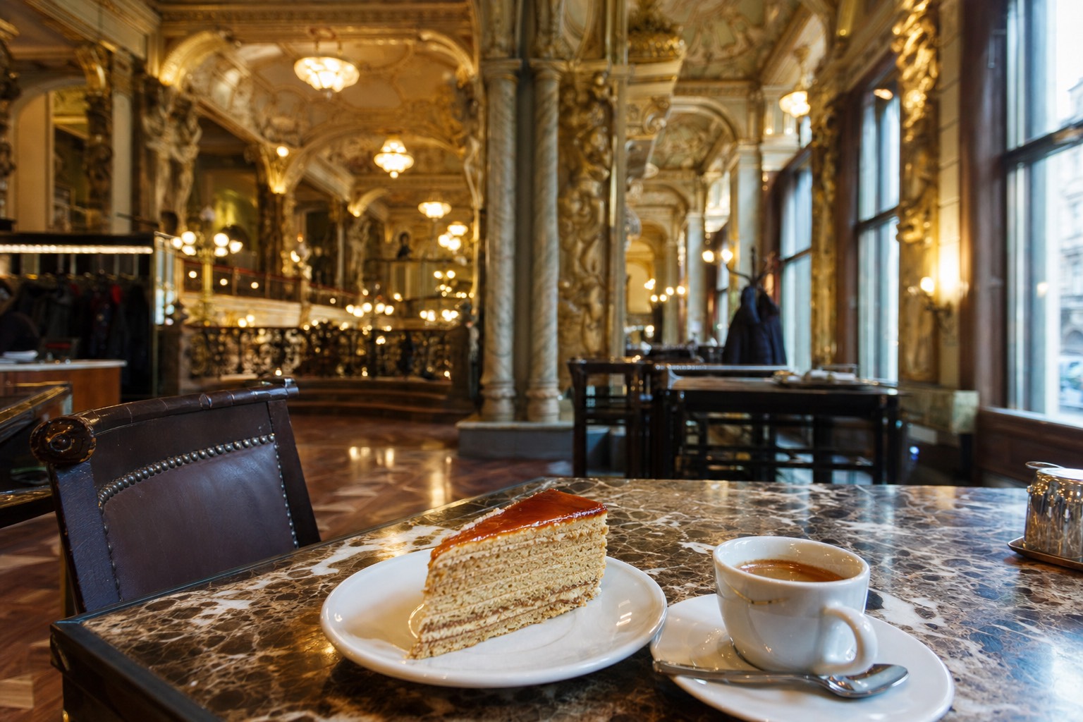 Marble café table holding a slice of layered Dobos cake with glossy caramel top and a small espresso in a porcelain cup, gilded columns and crystal chandeliers of a grand Budapest coffeehouse softly blurred behind, tall windows spilling morning light across parquet, old-world elegance, no people prominent