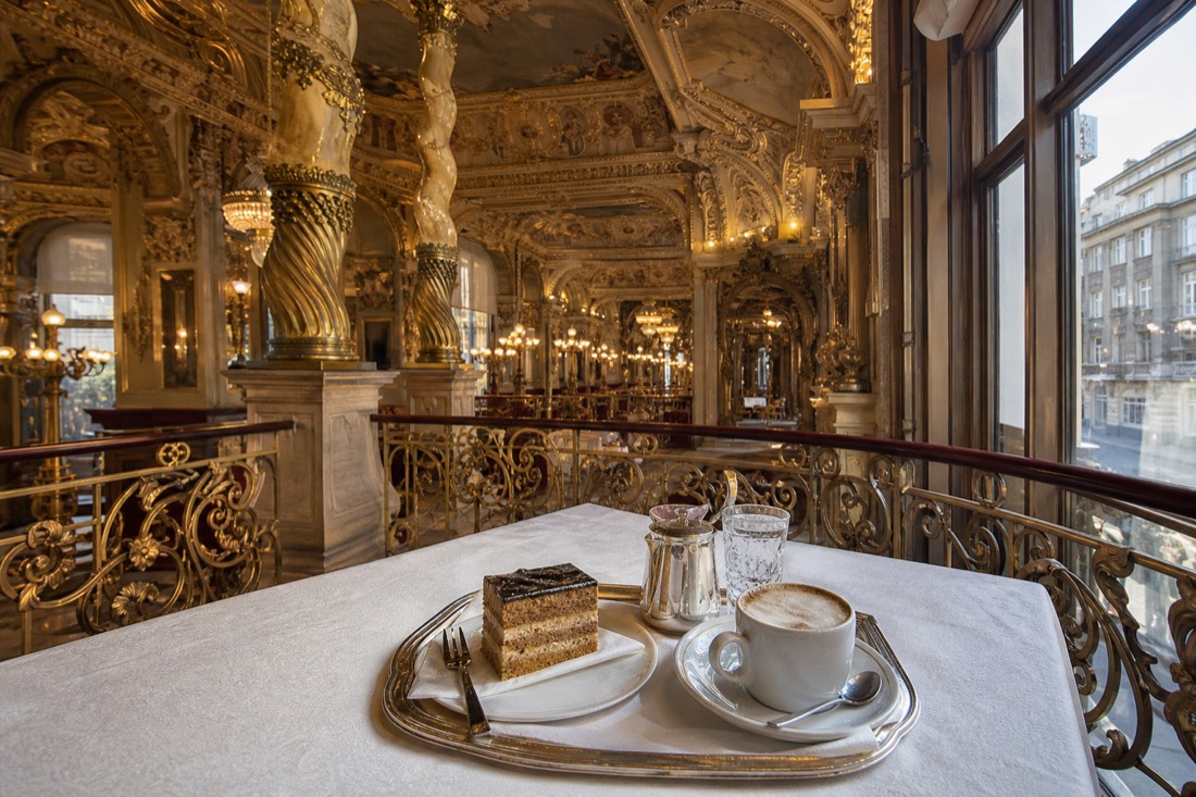 Interior of a historic Budapest coffeehouse with soaring frescoed ceiling, gilded twisted columns and marble tables, silver tray with coffee and layered cake on white linen in the foreground, warm chandelier glow against late-afternoon window light, wide view with no people in focus